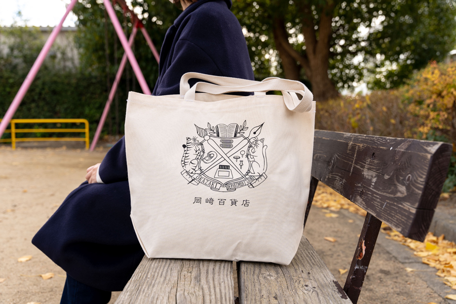 岡﨑百貨店のエンブレムがシルクスクリーンでプリントされた布バッグが冬の公園のベンチに置かれている