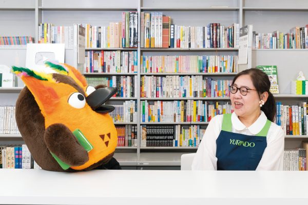 YouTube撮影スタジオのテーブルに並んだ岡﨑バイヤーとブッコロー（実写）、ブッコローが大笑いしている
