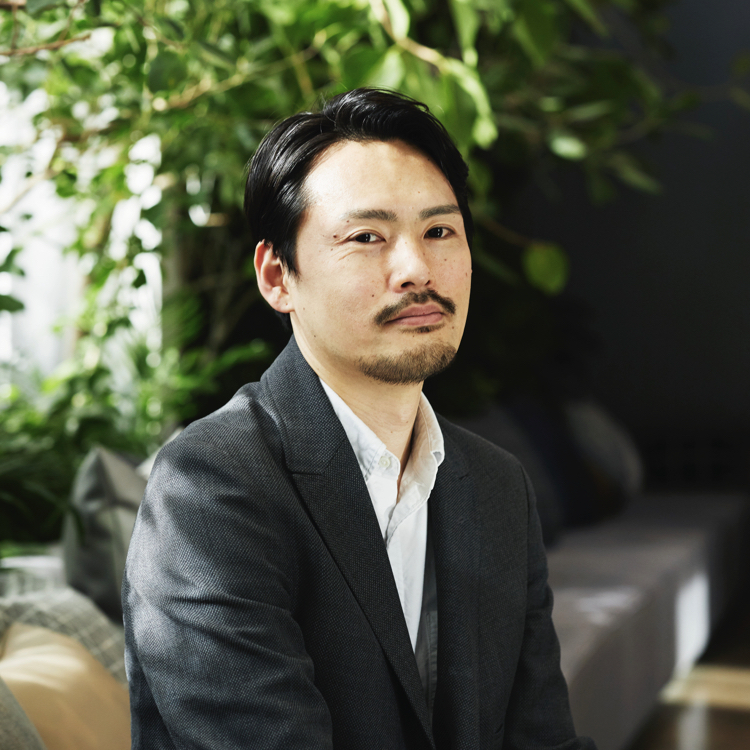 植物を背景に座る、山下正太郎さん近影。スーツにおひげ