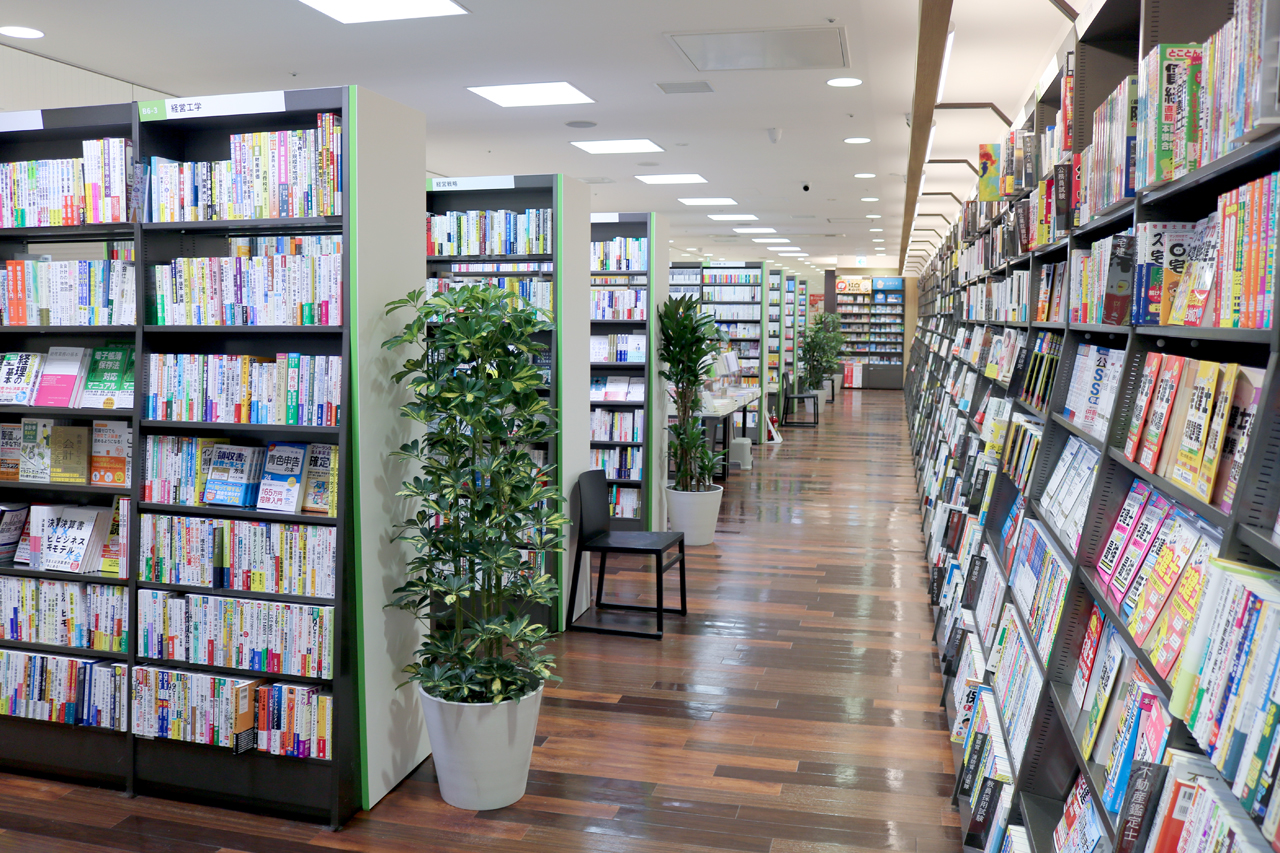 書籍売場、専門書コーナー。木目調の床に背の高い書棚、観葉植物