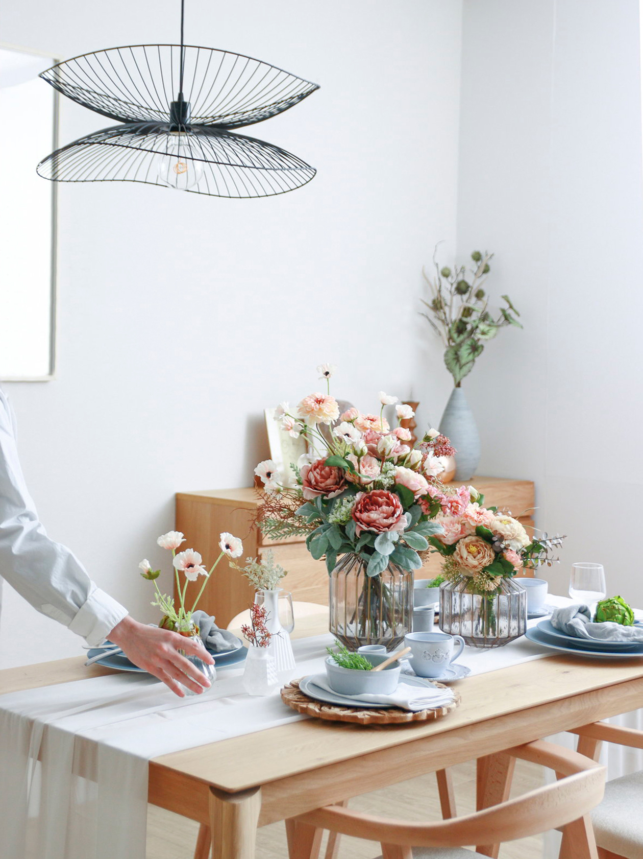 朝の食卓に造花とフェイクグリーンを飾ったイメージ。自然な木製のテーブル・椅子のセット、テーブルセンターの上に花瓶と精巧な造花の束。