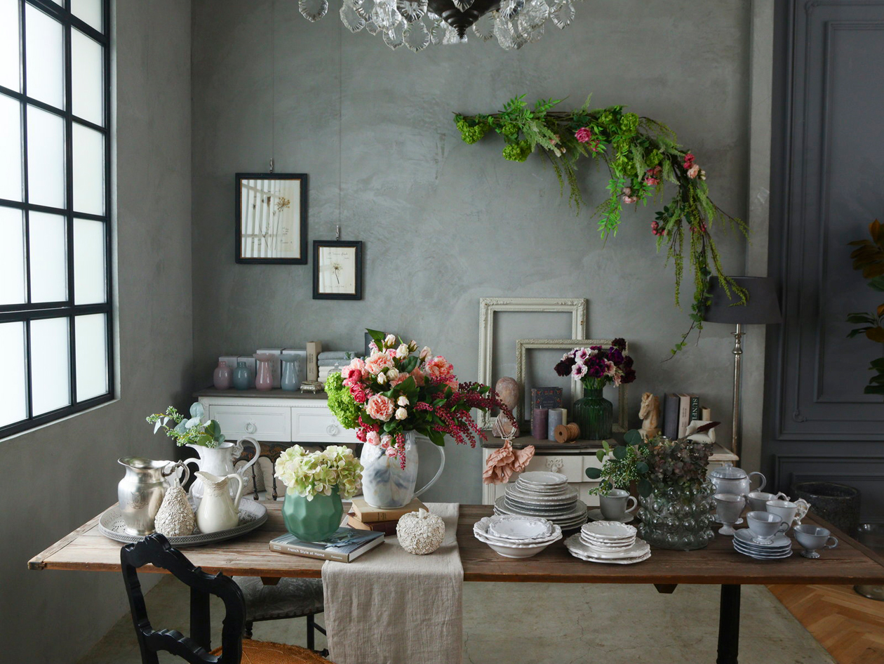 欧州風のシックな室内に造花とフェイクグリーンをセッティングしたイメージ。落ち着いた風合いの壁にフェイクグリーンのリース、花瓶に華やかな花束が差してある。