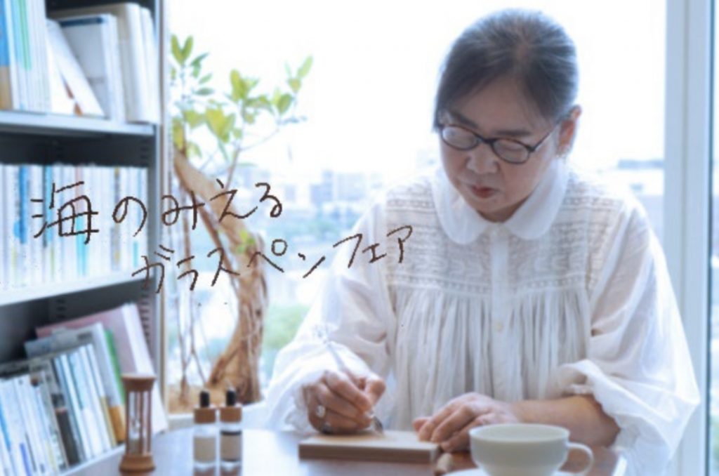 ストヨコの書棚と、その前でガラスペンを使って何か書いているバイヤー岡﨑。大きな窓から明るい光がさしている。
