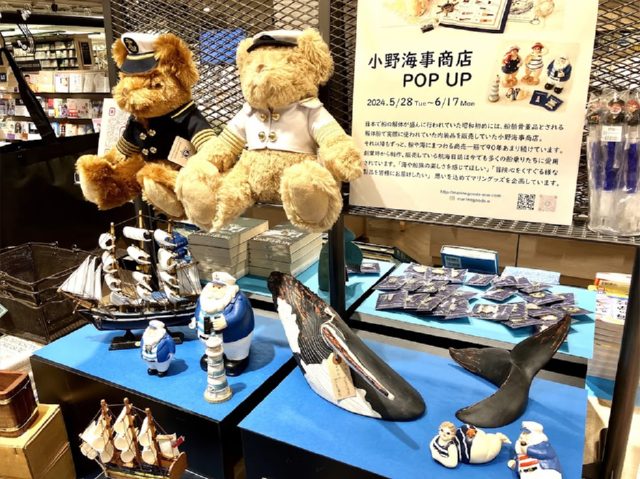 マリンルックのシャツを着たテディベアと、小野海事商店のパネル、クジラや船のホームデコレーションアイテムが海の色をした台の上に並んでいる
